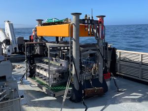A large scientific instrument sits on the silver deck of a ship on a sunny day. The instrument has a silver metal rectangular frame with an orange float at the top and several clear plastic chambers on the side. In the background is blue ocean with clear blue sky on the horizon.