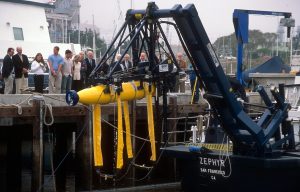 MBARI staff and Board of Directors observe crane operations for transport of a cylindrical autonomous robot with a yellow body and blue tail cone. The MBARI staff and members of MBARI’s Board of Directors are standing on the gray concrete pier. A blue metal crane is lifting the autonomous robot in the air at eye level with the people on the pier. Two yellow straps are hanging below the robot. The background is Moss Landing Harbor, with several other ships in the distance and overcast gray sky overhead.