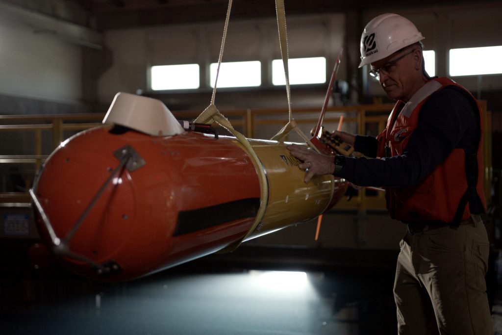 An MBARI engineer in an orange life vest, dark navy shirt, and brown pants wearing glasses and a white hard hat inspects an orange-and-yellow-colored, torpedo-shaped, autonomous robot. The engineer is holding a yellow control switch for an overhead crane. The robot is suspended from two yellow nylon straps. A large test tank is visible in the background.