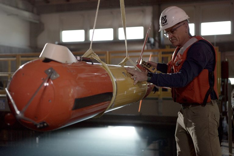 An MBARI engineer in an orange life vest, dark navy shirt, and brown pants wearing glasses and a white hard hat inspects an orange-and-yellow-colored, torpedo-shaped, autonomous robot. The engineer is holding a yellow control switch for an overhead crane. The robot is suspended from two yellow nylon straps. A large test tank is in the background.