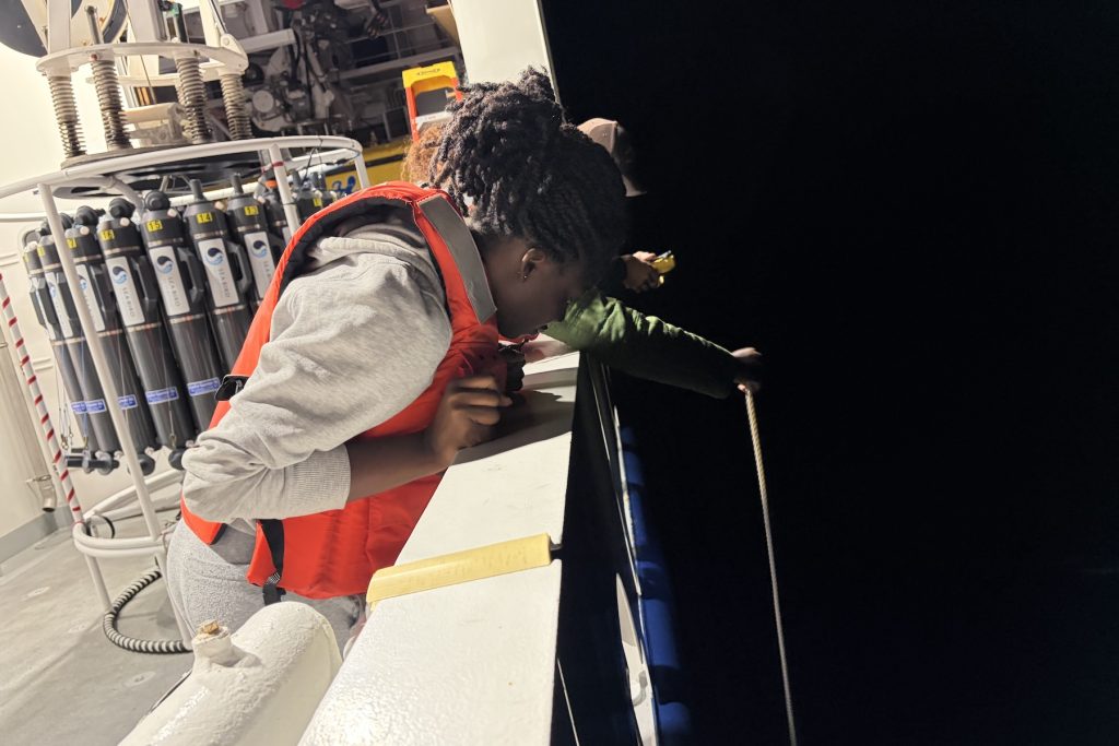 A university student wearing an orange life vest leans on the railing of a research ship to observe a trawl net collecting plankton at night.
