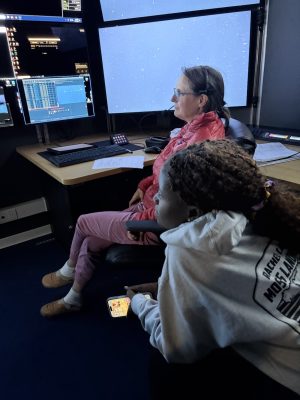 A scientist and a university student sit in the dark submersible control room aboard a research ship watching monitors displaying the live video feed from a robotic submersible exploring the deep sea.