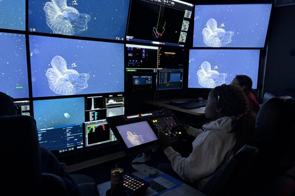 A university student sits in the dark submersible control room aboard a research ship. She is operating camera controls while watching a wall of monitors displaying the live video feed from a robotic submersible exploring the deep sea.