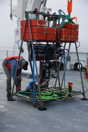 An engineer wearing an orange life vest checks cables on a scientific instrument on the back deck of a research ship on an overcast day.