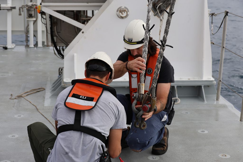 Two marine operations crew members prepare equipment on the deck of a research ship. Both are wearing white hard hats and orange life vests. The crew member on the right is holding two large metal clamps connected to thick gray ropes and a crane out of frame. In the background are maritime equipment, the silver metal deck of a research ship, and gray ocean.