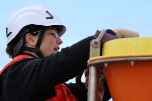 A scientist wearing a white hard hat and orange life vest adjusts the metal clasp securing a yellow lid on an orange funnel on a scientific instrument on the deck.