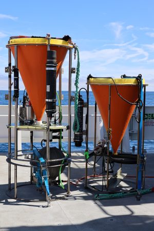wo scientific instruments sit on the deck of a research ship. The instrument on the left has an orange funnel with a yellow lid above a round silver metal frame with a silver metal sphere. The instrument on the right has an orange funnel with a yellow lid above a round silver metal frame. In the background is the gray metal deck of a research ship, with blue ocean and cloudy blue sky on the horizon.