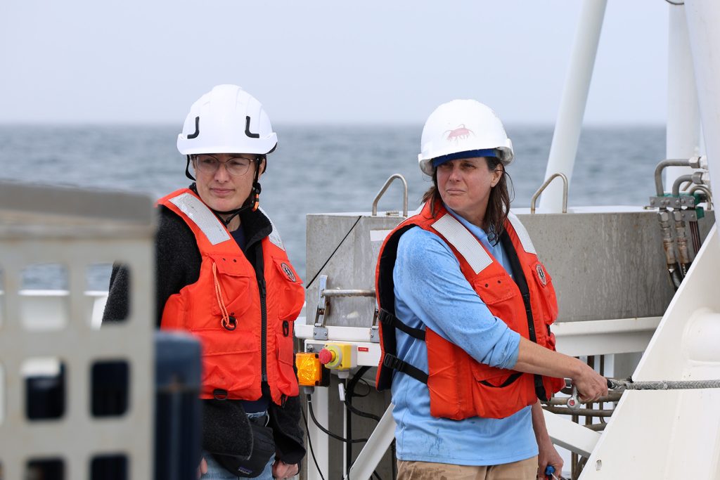 Two scientists observe preparations for deployment of scientific instruments on the back deck of a research ship. The scientists are wearing white hard hats and orange life vests. The scientist on the right is holding a gray rope. In the foreground is the blurred frame of a scientific instrument. In the background are maritime equipment, gray ocean, and overcast gray sky.