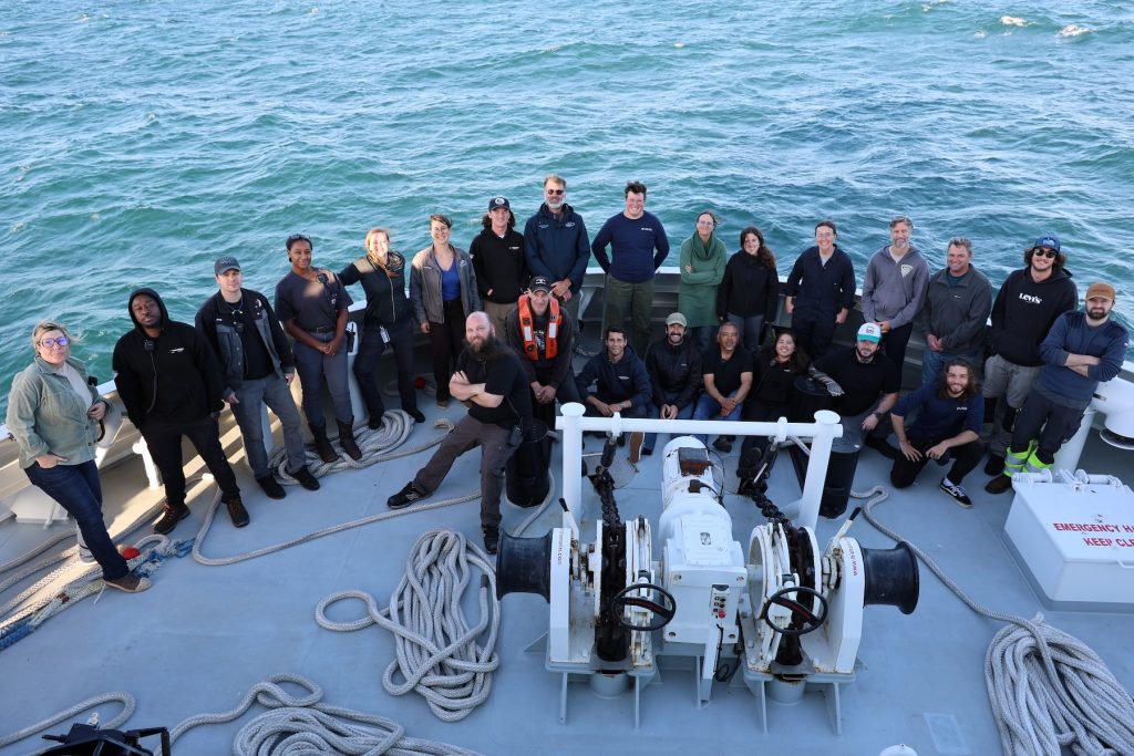 Twenty-four members of a scientific expedition pose for a photo on the bow of a research ship. In the foreground is maritime equipment. In the background is green-blue ocean.