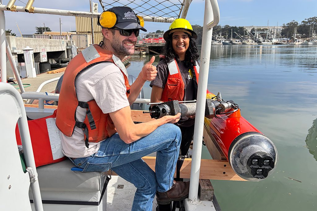 Two marine operations crew wearing life vests pose for a photo on a small boat while testing a sensor next to an autonomous robot. The robot has an orange-and-yellow cylindrical plastic housing and a black camera array.