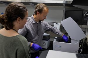 Two researchers prepare samples for genetic sequencing. The researcher on the left has brown hair in a loose bun and is wearing glasses and a dark green sweater. The researcher on the right has short blond hair and is wearing a brown-striped shirt and bright-purple nitrile gloves. On the right side of the frame is a gray DNA sequencing instrument. In the background are silver metal shelves and a white wall.