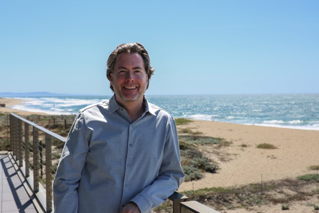 CeNCOOS Director Henry Ruhl poses for a portrait outdoors. He is wearing a light blue collared, button-down shirt and leaning against a silver metal railing. In the background are sand dunes, the beach, blue-green ocean with large white waves, and clear blue sky on the horizon.