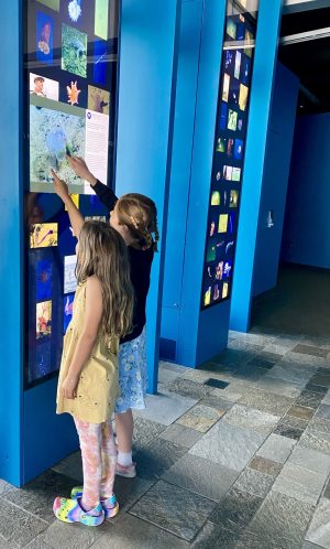 Two children point at the screen of an interactive museum exhibit. The child on the left has long blonde hair and is wearing a yellow dress, orange-and-peach patterned leggings, and rainbow-colored clogs. The child on the right has blonde hair in braids and is wearing a black sweatshirt, blue floral-print skirt, white socks, and pink shoes. The interactive exhibit has a bright-blue wooden frame and a tall vertical video screen displaying tiles of photos with one large photo in the middle with a white box and black text. The background includes a second bright-blue interactive kiosk, a bright-blue wall, gray carpet, and gray stone floor tiles.