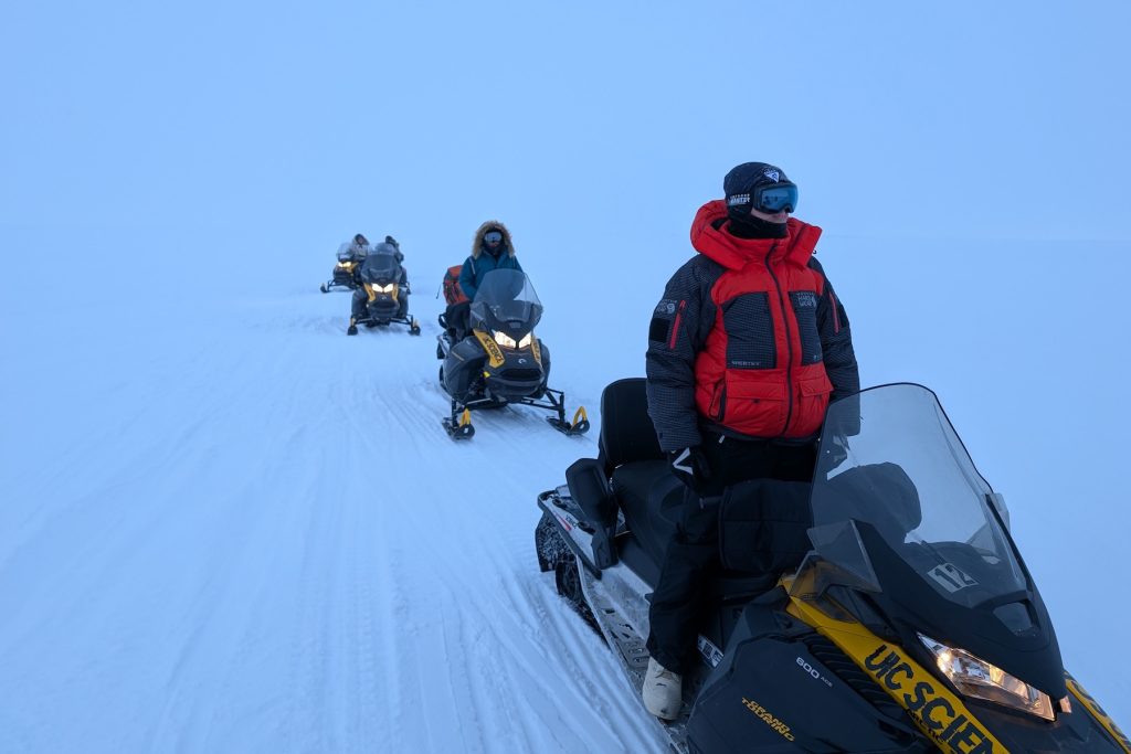 Four members of an expedition team ride snowmobiles to a research site on Arctic sea ice. The expedition team members are wearing winter jackets and goggles and each is riding their own snowmobile. The background is white ice and white sky.