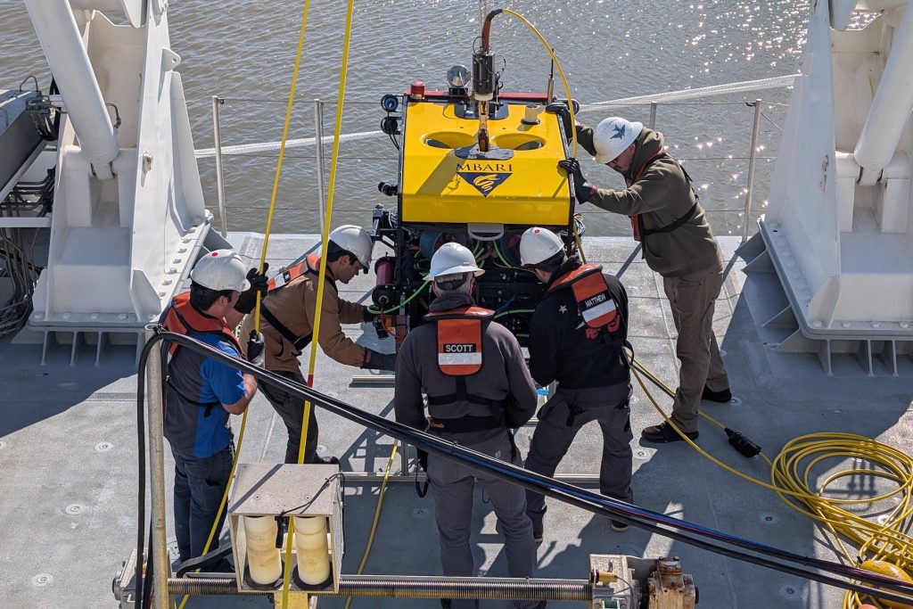 Five marine operations crew members prepare a robotic submersible for a test deployment from the deck of a research ship on a sunny day. The crew members are wearing white hard hats and orange life vests. The robotic submersible has a yellow float with the MBARI logo, a black metal frame, and assorted scientific instruments. It is sitting on a silver metal frame and connected to a crane by a yellow tether cable. In the foreground is assorted maritime equipment. In the background is the brown water of a harbor.