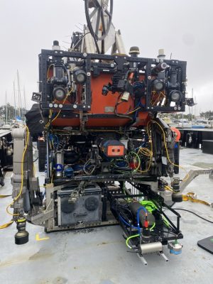A robotic submersible sits on the outdoor deck of a research ship docked in a harbor. The submersible has an open drawer-like metal sled of instruments for seafloor mapping.