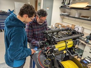 Two engineers adjusting electronics on an underwater robot in the lab. The engineer on the left is wearing glasses and a fuzzy blue hooded fleece sweatshirt. The engineer on the right is wearing glasses and a long-sleeved shirt with a blue, red, and gray plaid print. The robot in the foreground has a black metal frame surrounding a yellow metal cylinder with brightly colored cables extending from the left side. The background is a white laboratory with shelves of engineering equipment.