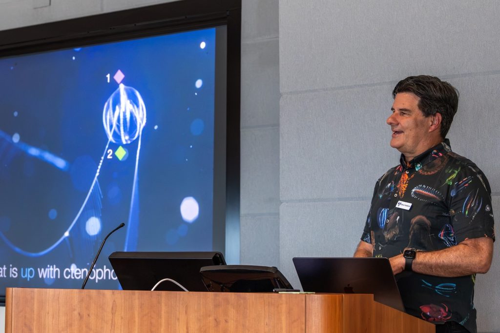 A scientist speaks at a scientific workshop. He is standing behind a wooden podium in front of a projector screen displaying a slide with a photo of a comb jelly captioned, What is up with ctenophores?