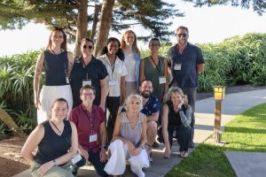 Eleven workshop participants pose for a photo outdoors on a sunny day in front of a hedge of leafy green plants.