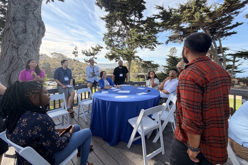 Ten workshop participants participate in a discussion outdoors on a sunny day. Some participants are seated, and others are standing.