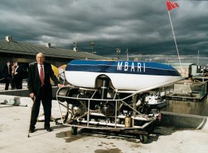 MBARI’s founder David Packard, wearing glasses, a black suit, white shirt, and red tie and holding a bronze cane, stands on MBARI’s gray concrete dock next to a deep-sea robot. The deep-sea robot has a blue-and-white float and silver metal frame, and is on a wooden dolly. In the background is a gray building with a gray roof, Moss Landing Harbor, and a cloudy gray sky.