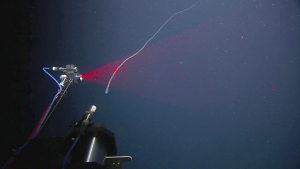 An imaging instrument scans a rope-like deep-sea siphonophore with a sheet of red laser light. The instrument is mounted to a robotic submersible outside of the frame. The instrument is in a silver metal cylinder housing attached to the end of a black metal rod extending from the left corner of the frame. Blue and red cables connect the instrument to the submersible. At the bottom of the frame is a black cylindrical camera housing mounted to the submersible. In the background is dark blue water dotted with scattered white flecks of organic material.