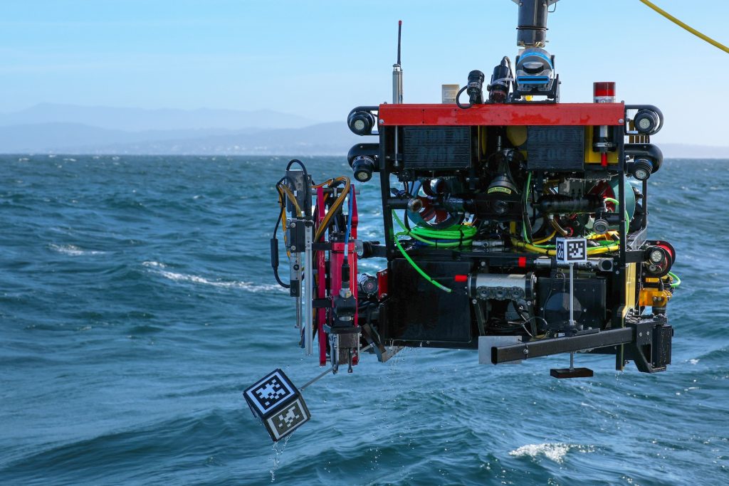 A robotic submersible returns from deployment in the ocean. The robotic submersible has a red float, a black metal frame, a red robotic arm, a variety of multi-colored cables, and various scientific instruments. The submersible is suspended from a tether and crane out of frame. In the background is choppy greenish-blue ocean with coastal mountains and blue sky on the horizon.