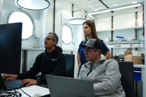 An engineer and two scientists review data on computers in a lab aboard a research ship. In the background are the white walls and shelves of the lab and porthole windows looking out to sea.