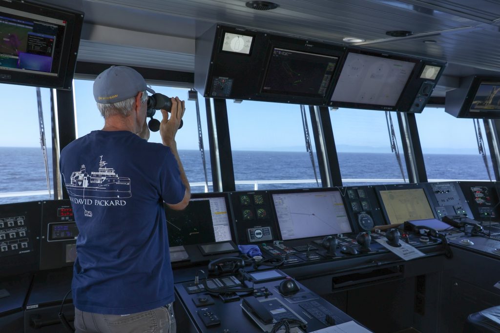 A marine operations crew member looks out to sea from the bridge of a research ship. He is standing in front of a counter of ship controls and using binoculars to look out large windows. In the background is blue ocean, coastal mountains, and blue sky on the horizon.