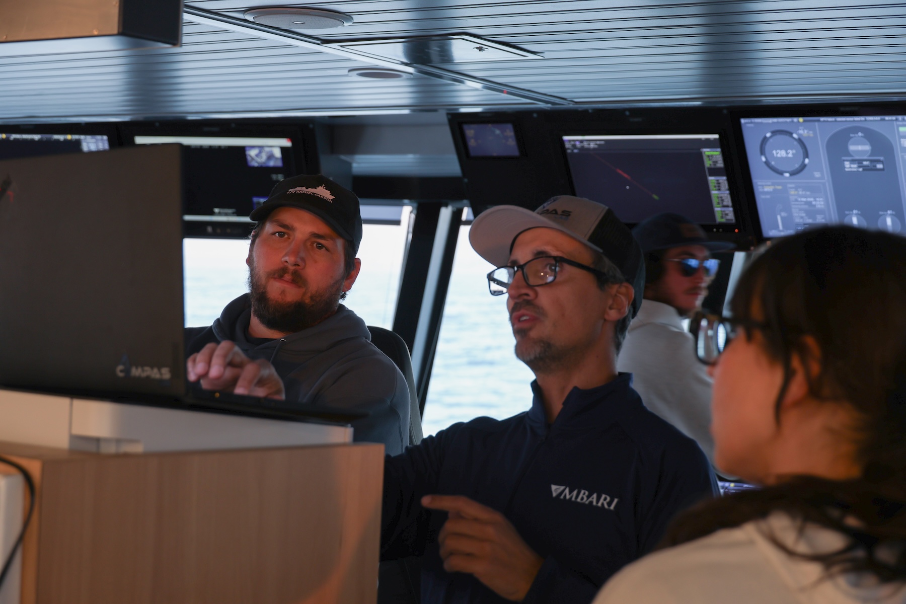 Three members of the expedition team discuss plans for the next day’s dives in the bridge of a research ship. The engineer in the center is using a laptop computer to show data to the marine operations crew. In the background are monitors with maritime data, a member of the ship’s crew, and large windows looking out to the ocean.