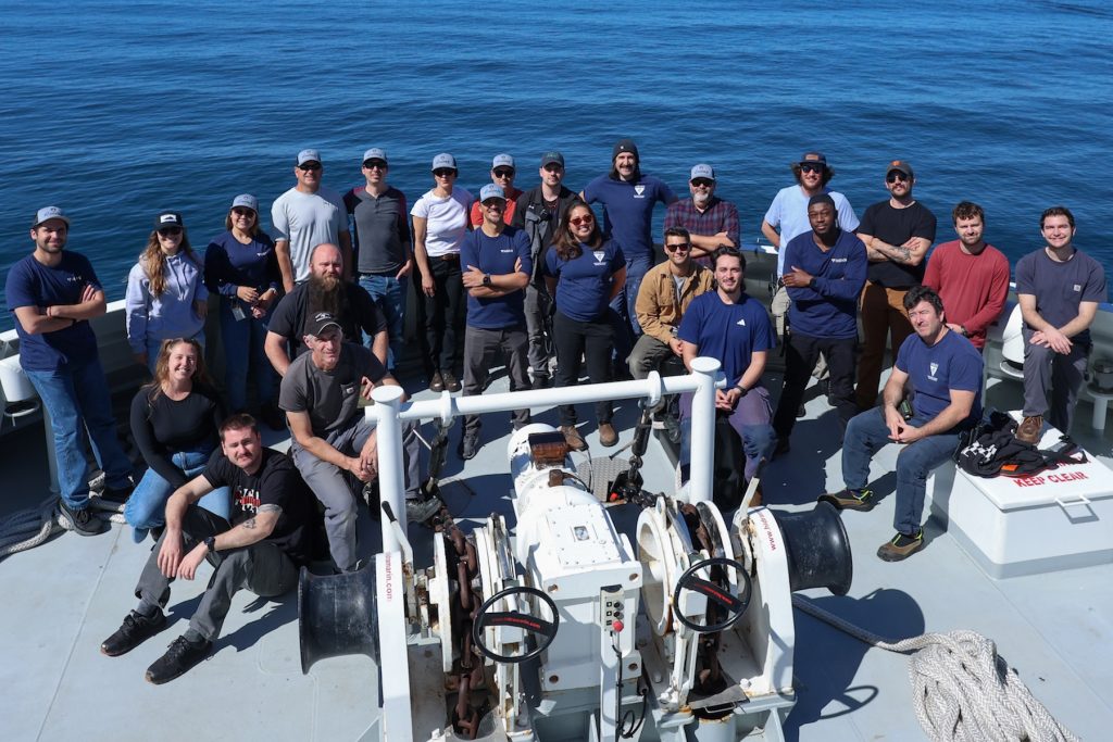 Twenty-four members of a scientific expedition pose for a photo on the bow of a research ship. In the foreground is maritime equipment. In the background is blue ocean.