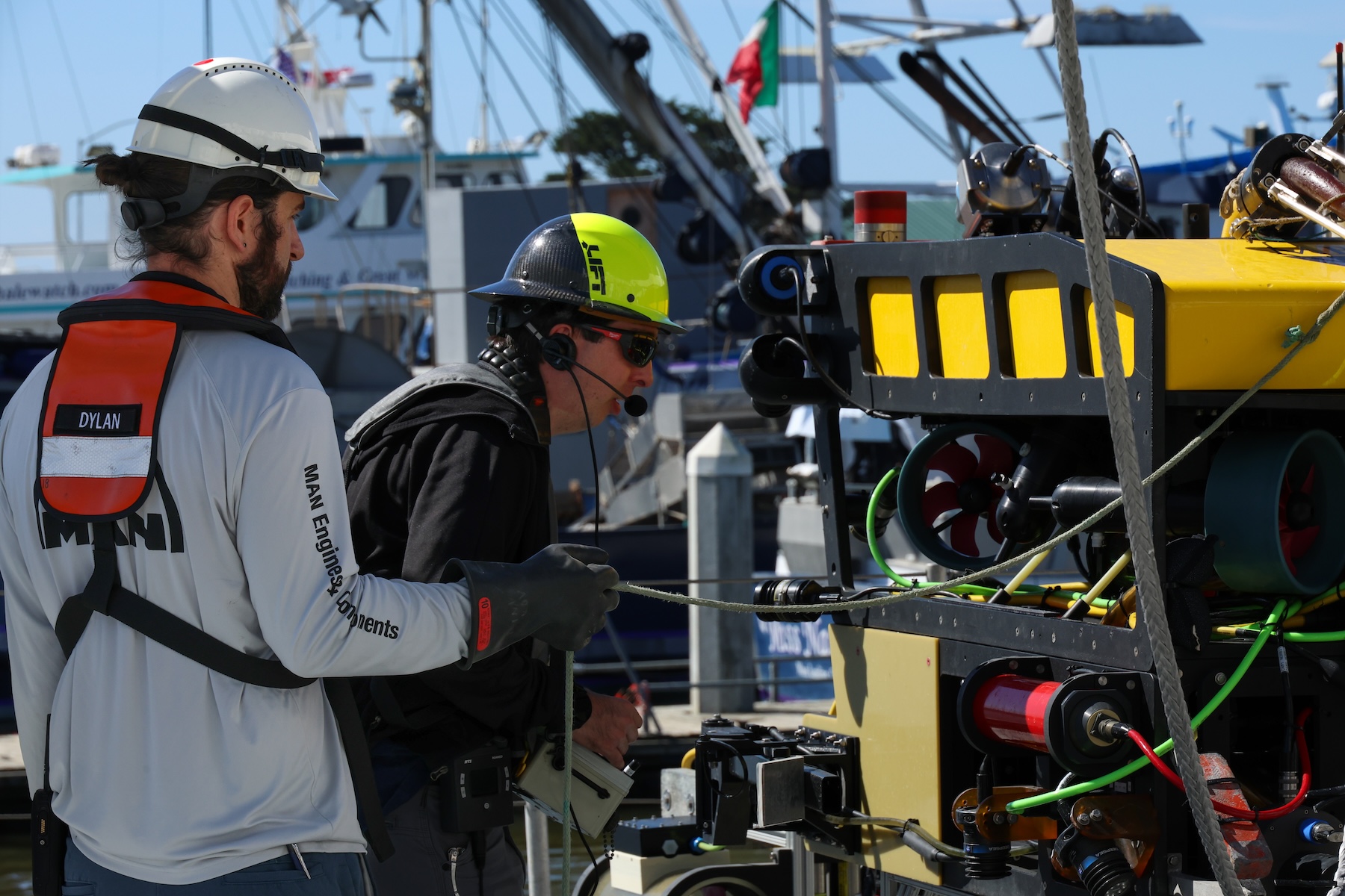 Two marine operations crew members inspect a robotic submersible on the deck of a research ship on a sunny day. The crew members are wearing hard hats and orange life vests. The submersible has a yellow plastic float, black metal frame, and multi-colored cables and instruments. In the background are neighboring ships in the harbor, maritime equipment, and bright blue sky.