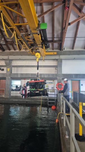 A mechanical engineering technician assists with deployment of the robotic submersible in an indoor test tank. The technician is wearing a white hard hat, gray sweatshirt, and blue jeans, and holding controls for a crane while standing against the railing of a large pool of water. The submersible has a black metal frame with multi-colored cables and instruments and is suspended from a yellow crane overhead.