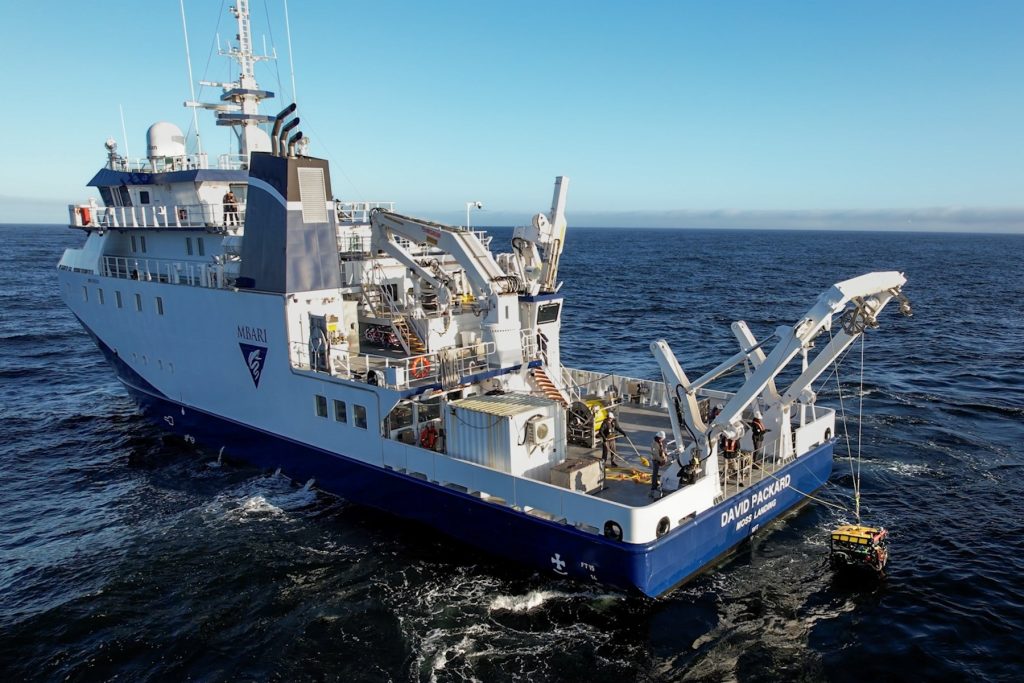 An aerial drone photo shows the deployment of a robotic submersible from the back deck of a research ship. The submersible has a yellow float and black metal frame, and is suspended from a crane by several tether lines. The research ship is white with a blue hull with the MBARI logo printed on the side and the name and location, David Packard and Moss Landing, printed on the stern. The background is blue ocean with coastal mountains and blue sky on the horizon.
