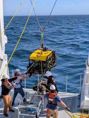 Four marine operations crew members standing on the deck of a research ship recover a robotic submersible following its deployment at sea. The crew members are wearing white hard hats and orange life vests. The submersible has a yellow float, black metal frame, and multi-colored cables and electronic equipment. The background is green-blue ocean with blue sky on the horizon.