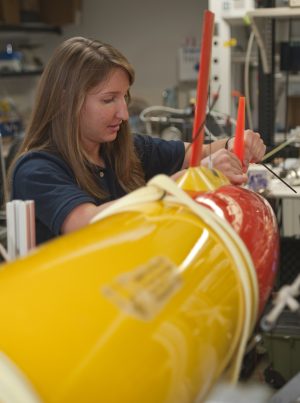 An MBARI summer intern with long brown hair wearing a blue shirt adjusting fins on an orange-and-yellow-colored, torpedo-shaped, autonomous robot. Various lab equipment is visible in the background.