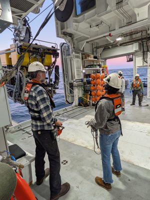 Four people in life vests and hard hats stand in a hangar aboard a research ship as a large robotic submersible is craned onboard.