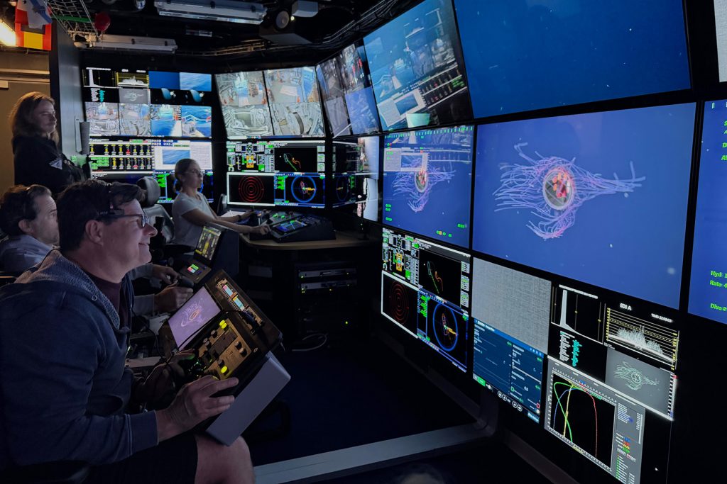 A scientist sits in the dark submersible control room aboard a research ship with three submersible pilots, watching a live video feed of a colorful deep-sea jelly on a wall of monitors.