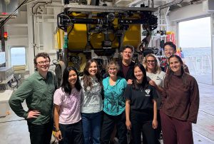 Nine smiling scientists pose for a photo inside a hangar aboard a research ship, in front of a large robotic submersible with a yellow float.