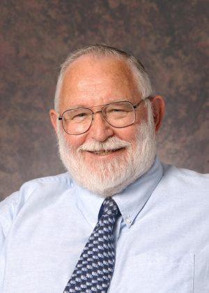 Portrait of Richard Barber wearing a pale blue shirt, patterned dark blue tie, and metal-frame glasses. This portrait was taken against a mottled mauve background.