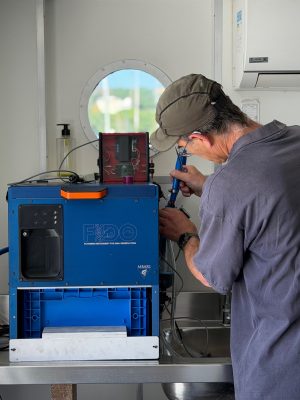 An engineer adjusts a scientific instrument inside a lab aboard a research ship. The engineer has his back to the camera. The instrument has a blue metal housing with the names and logos for FIDO and MBARI. In the background are the white metal walls of a research ship laboratory with a round porthole window.