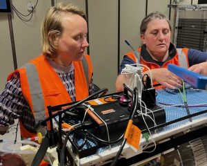 Two researchers wearing bright orange work vests prepare a scientific instrument in the lab aboard a research vessel. In the foreground is an MBARI Environmental Sample Processor instrument. In the background are beige walls of a scientific lab.