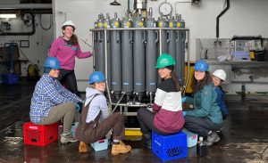 A researcher with curly brown hair wearing a white hard hat, pink pullover, and black pants stands to the left of a carousel of tall gray plastic water collection bottles. Five researchers seated on plastic crates around the carousel are collecting water samples. The background is the deck of an oceanographic research ship with assorted scientific instruments and maritime gear.