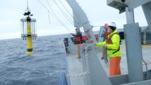 A researcher wearing a white hard hat, black beanie, bright yellow jacket, orange life vest, and orange pants stands on the deck of a research ship holding a rope line while deploying a yellow plastic sediment trap from a crane. The right side of the frame is the gray metal deck of a research ship, with two other researchers supporting deployment. On the left of the frame is gray-blue ocean with pale-blue sky on the horizon.