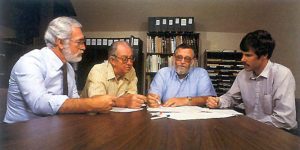 Four members of MBARI’s inaugural leadership team sitting around a brown table reviewing documents. In the background are bookcases full of binders and paper files.
