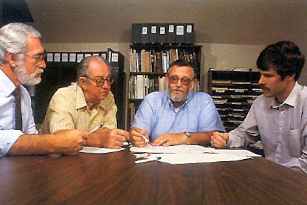 Four members of MBARI’s inaugural leadership team sitting around a brown table reviewing documents. In the background are bookcases full of binders and paper files.