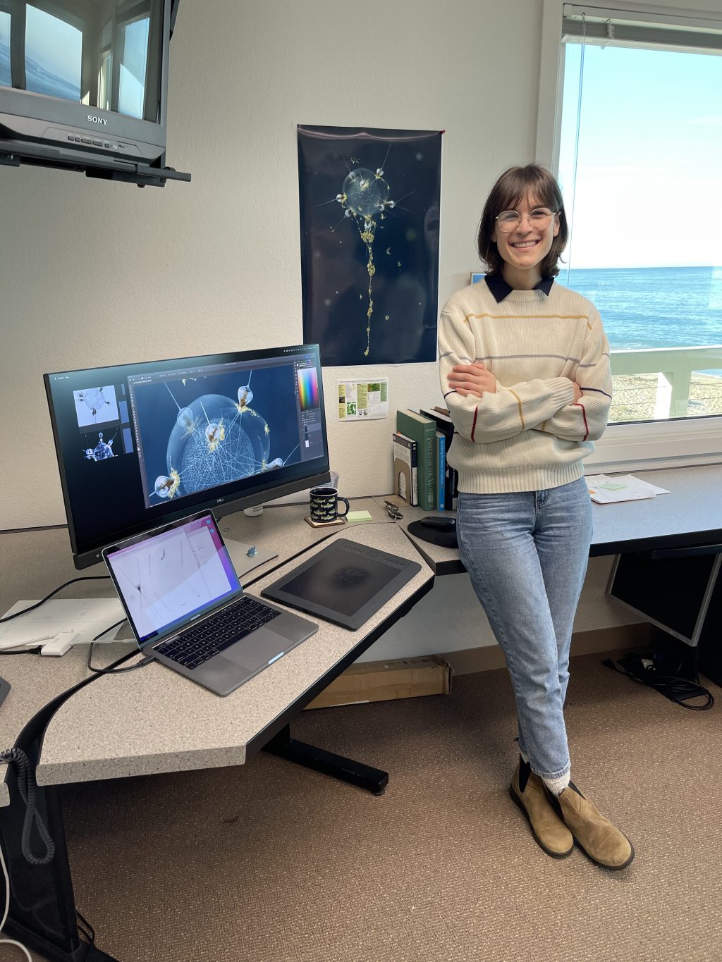 Emily Mitchell standing in her office space, including a computer screen with illustration editing software open and poster of phaeodarian illustration hanging behind her.