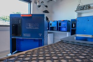 A scientific instrument sits in a research lab. The instrument has an orange handle, a bright blue plastic basket, and a square blue metal frame printed with the instrument’s name FIDO Filtering Instrument for DNA Observation, and logos for FIDO, USGS, and MBARI. Four additional instruments are lined up in a row in the background. In the foreground are several clear round plastic pucks with black rubber seals. In the background are a window and the white walls of a research lab.