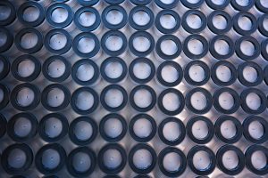 A tray of dozens of clear, round plastic pucks with black rubber rings waits for inspection.