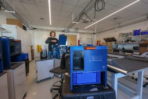 An engineer with shaggy blond hair wearing a black sweatshirt and blue jeans prepares a scientific instrument in a lab. He is adjusting the instrument’s blue metal frame. In the foreground is another instrument with a square blue metal frame printed with the name FIDO Filtering Instrument for DNA Observation and logos for FIDO, USGS, and MBARI. In the background are benches, shelves, and cabinets of assorted engineering equipment.